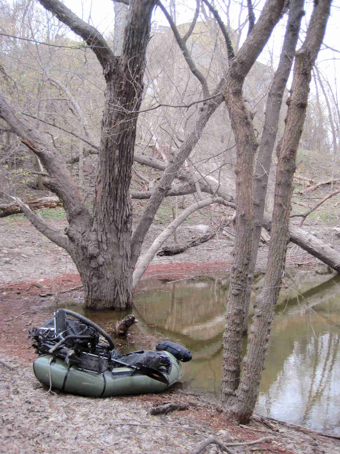 An olive green inflatable raft with a disassembled bike on top, on a dirt river bank in the bare woods